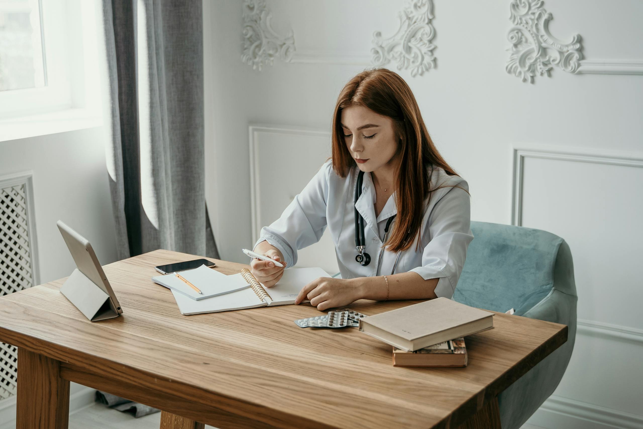 A female doctor in a lab coat writing notes at a wooden desk with a stethoscope and tablets nearby.